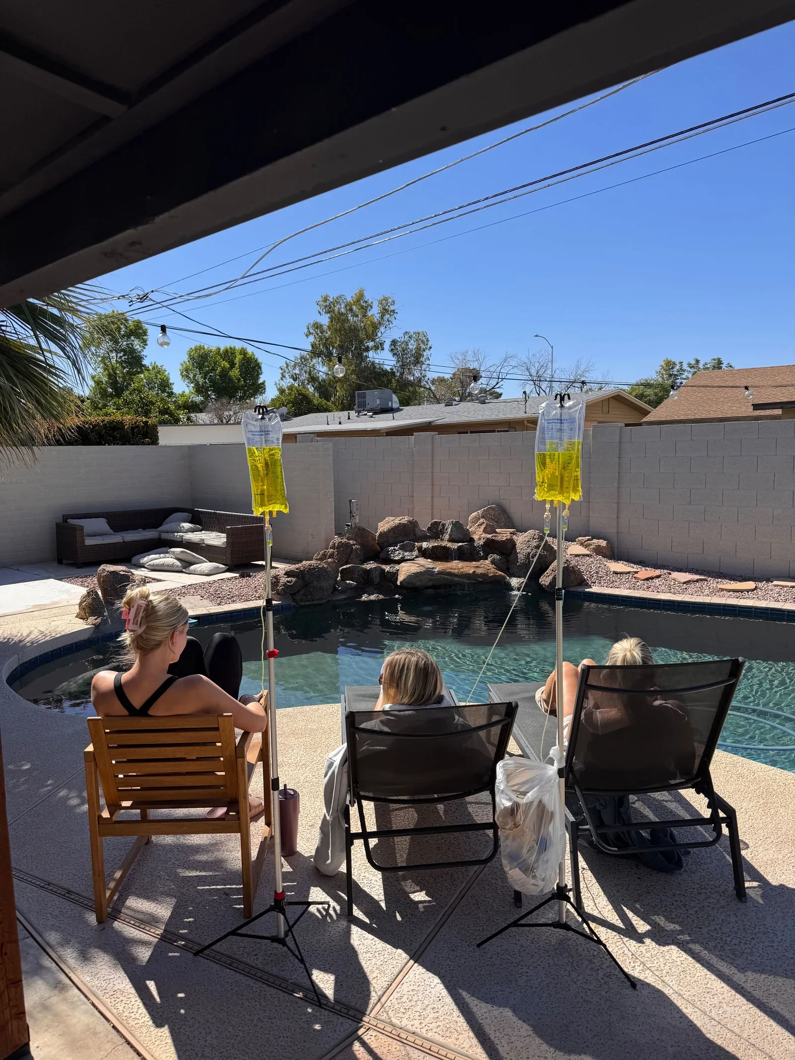 Three people receiving IV treatments on patio chairs beside a Phoenix pool, stands set up on the deck