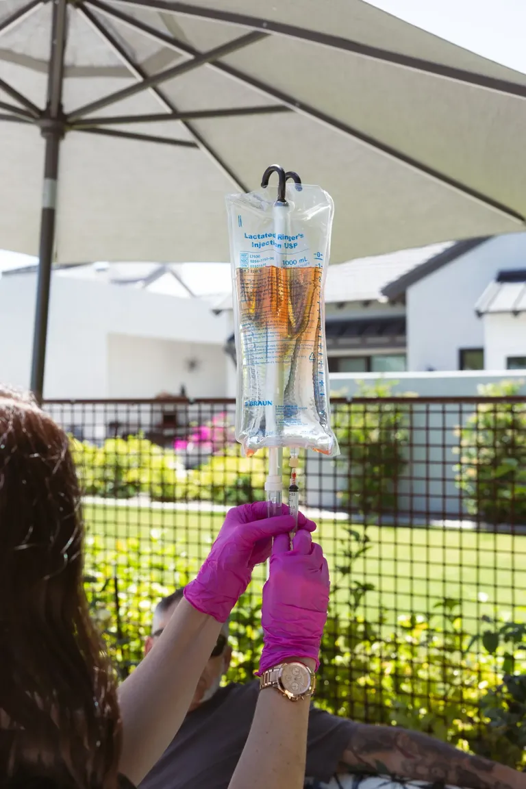 A nurse in pink gloves adjusting a hanging IV bag under a patio umbrella on a bright Phoenix afternoon